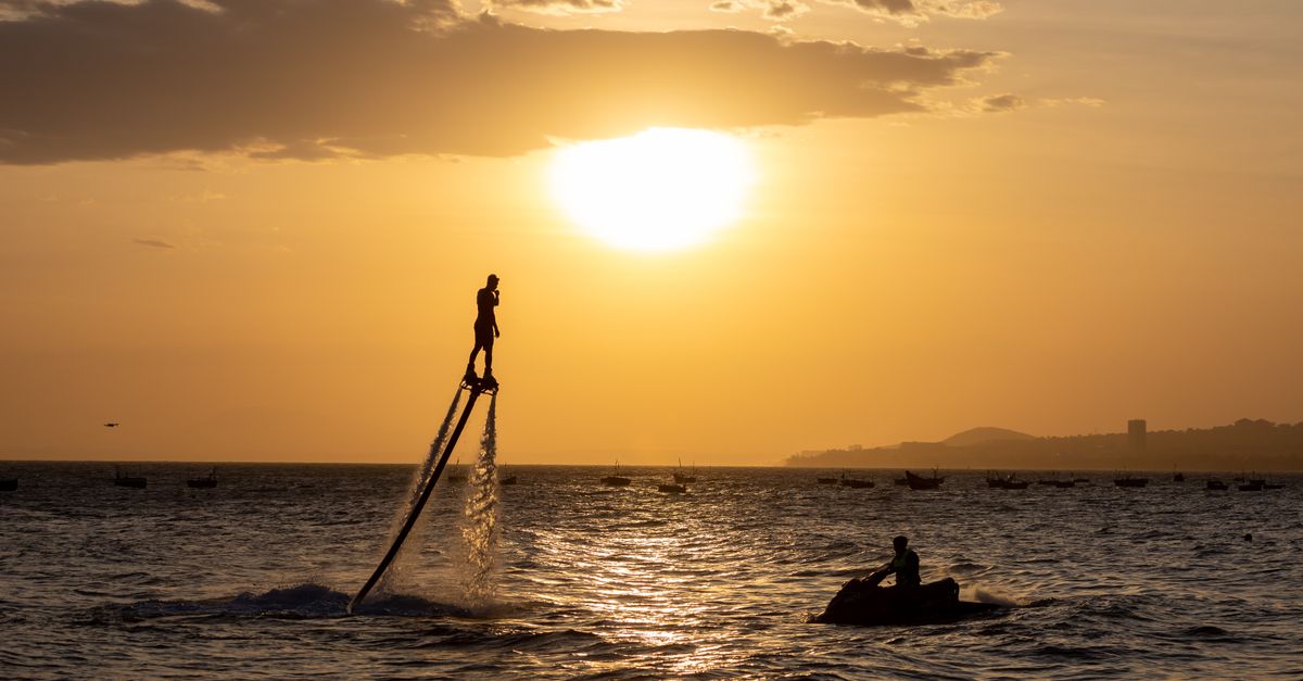 A person flyboarding at sunset over a vibrant sea with a jet ski nearby.