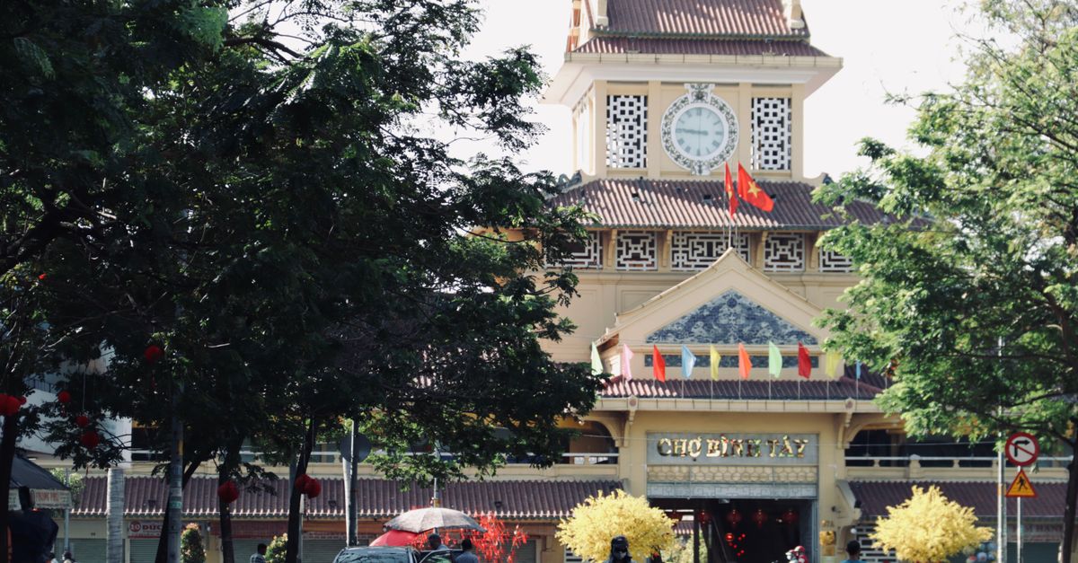 Scenic view of the iconic Binh Tay Market entrance with surrounding trees and vibrant flags in Ho Chi Minh City, Vietnam.