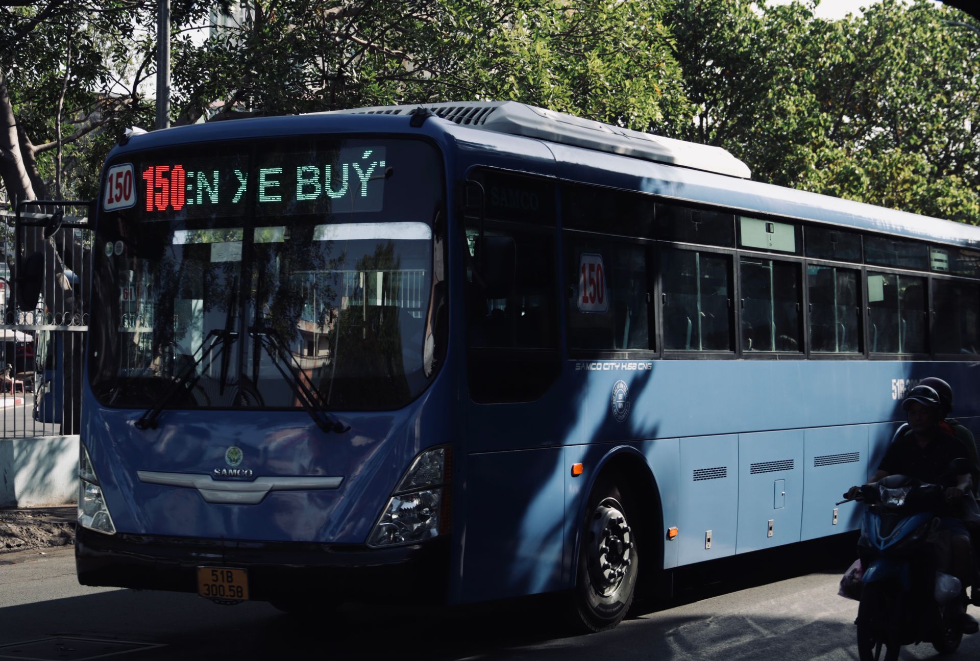 Blue bus and motor scooter on a sunlit street in Vietnam, capturing urban life and transportation.