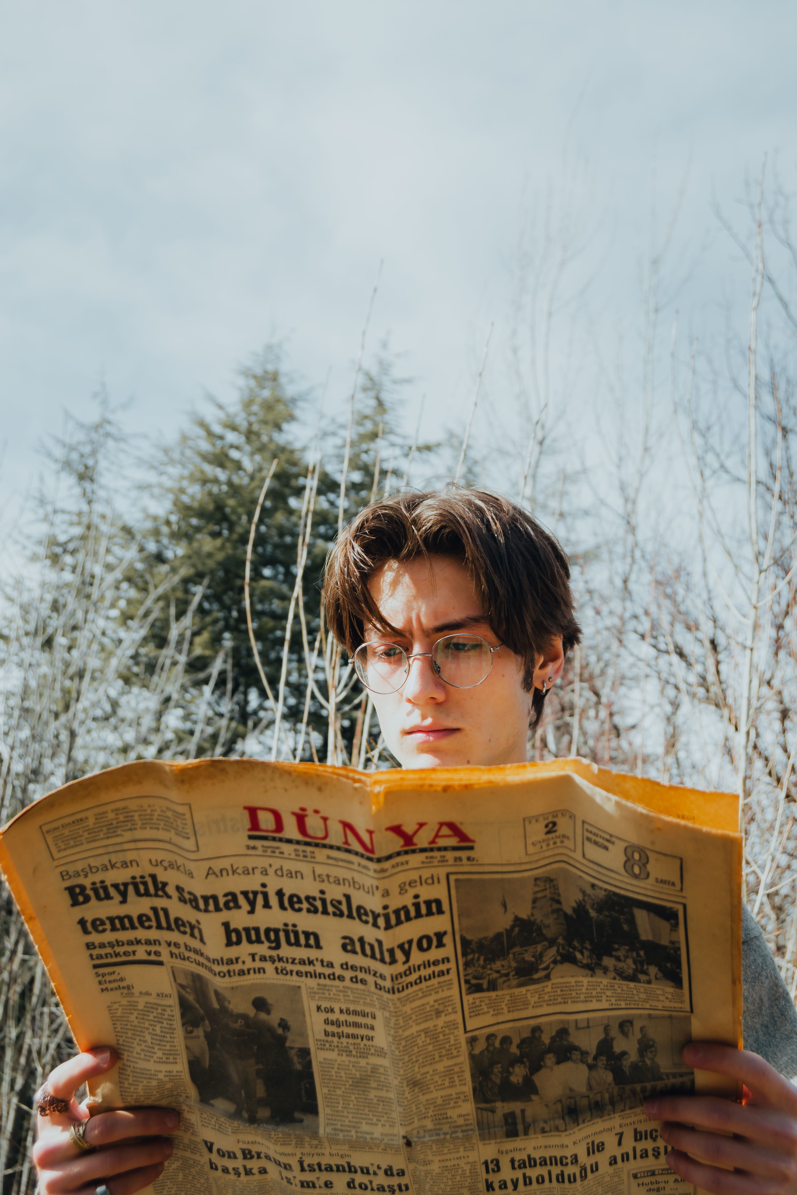 A young caucasian man with glasses reads a newspaper outside on a sunny day.
