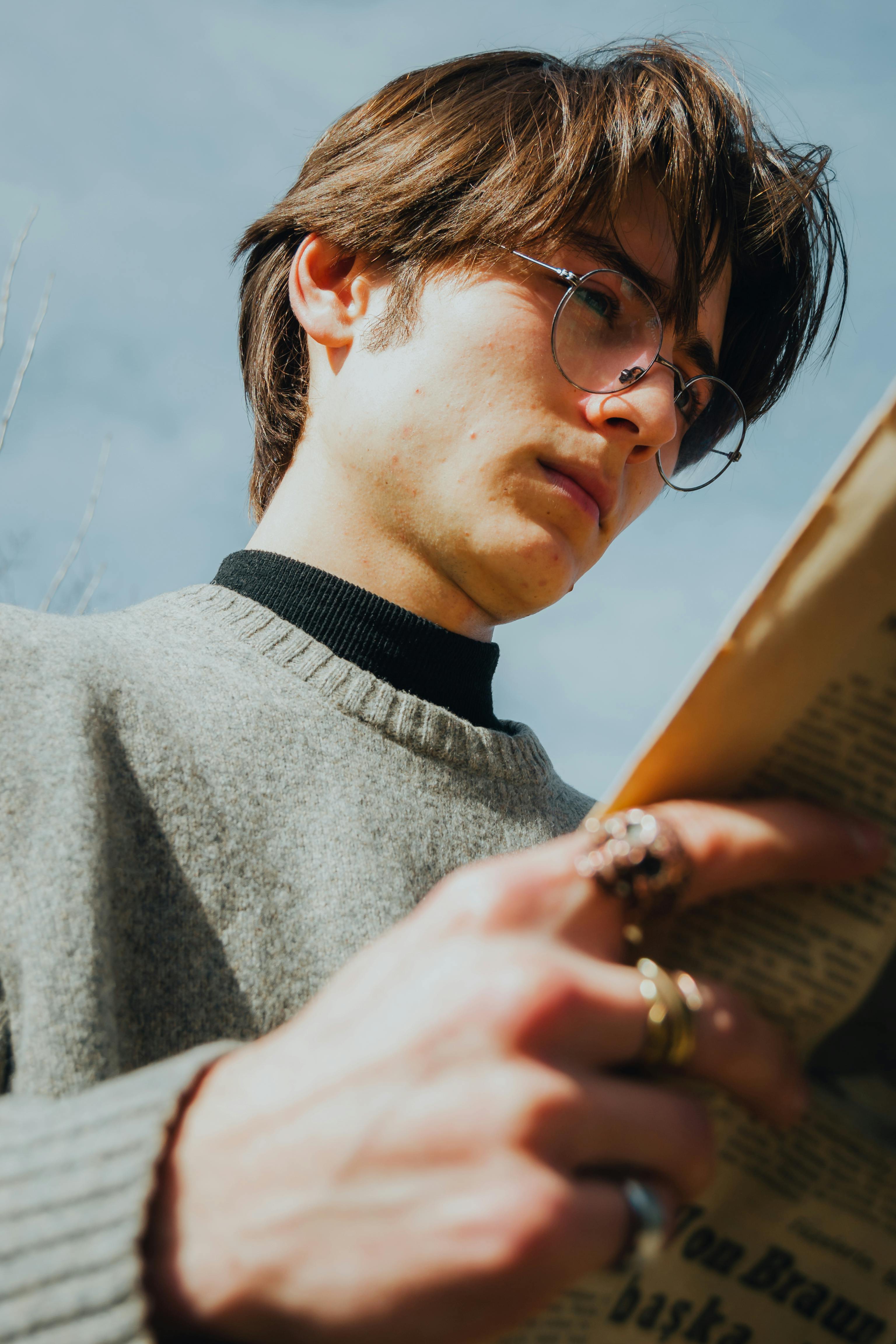 Portrait of a young man wearing eyeglasses and a sweater, intently reading a newspaper outdoors.