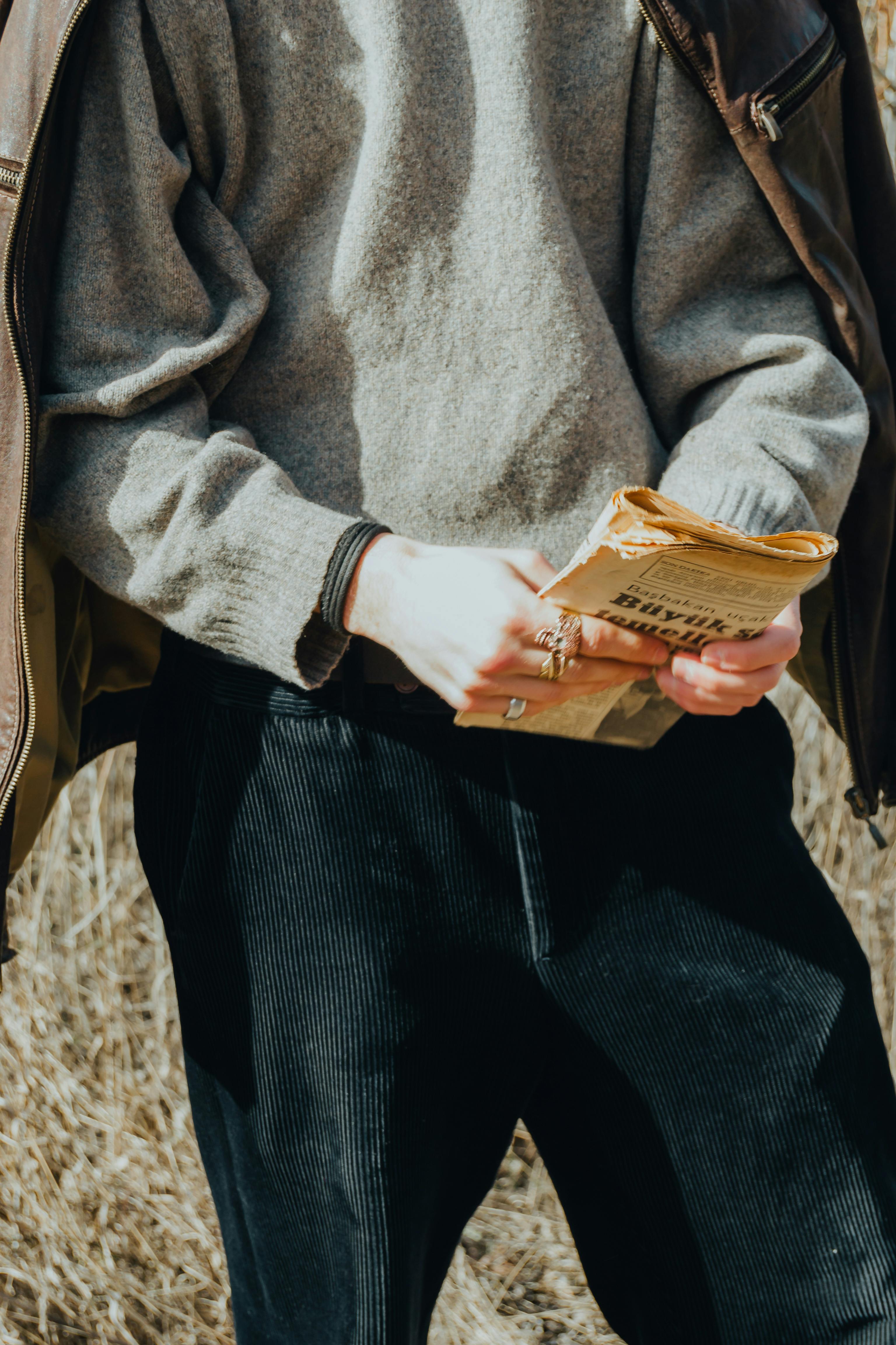 A person in a sweater holds an old newspaper outdoors, showcasing casual fashion style.