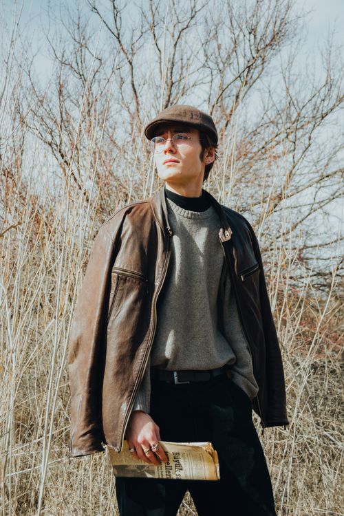 Free Young man in fashionable attire with a leather jacket and cap standing outdoors holding a newspaper. Stock Photo