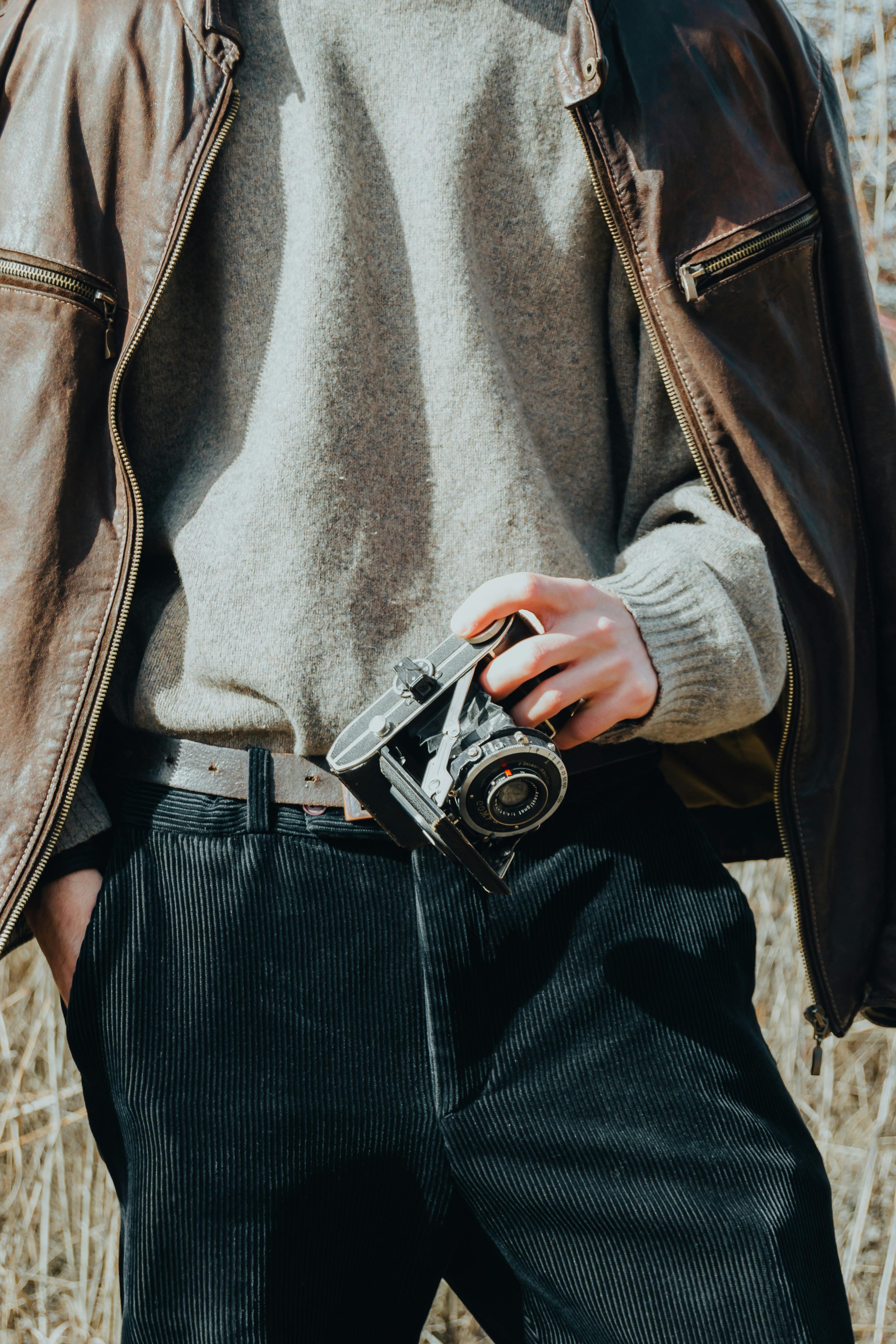 A person holding a vintage analog camera, wearing a leather jacket and sweater outdoors.