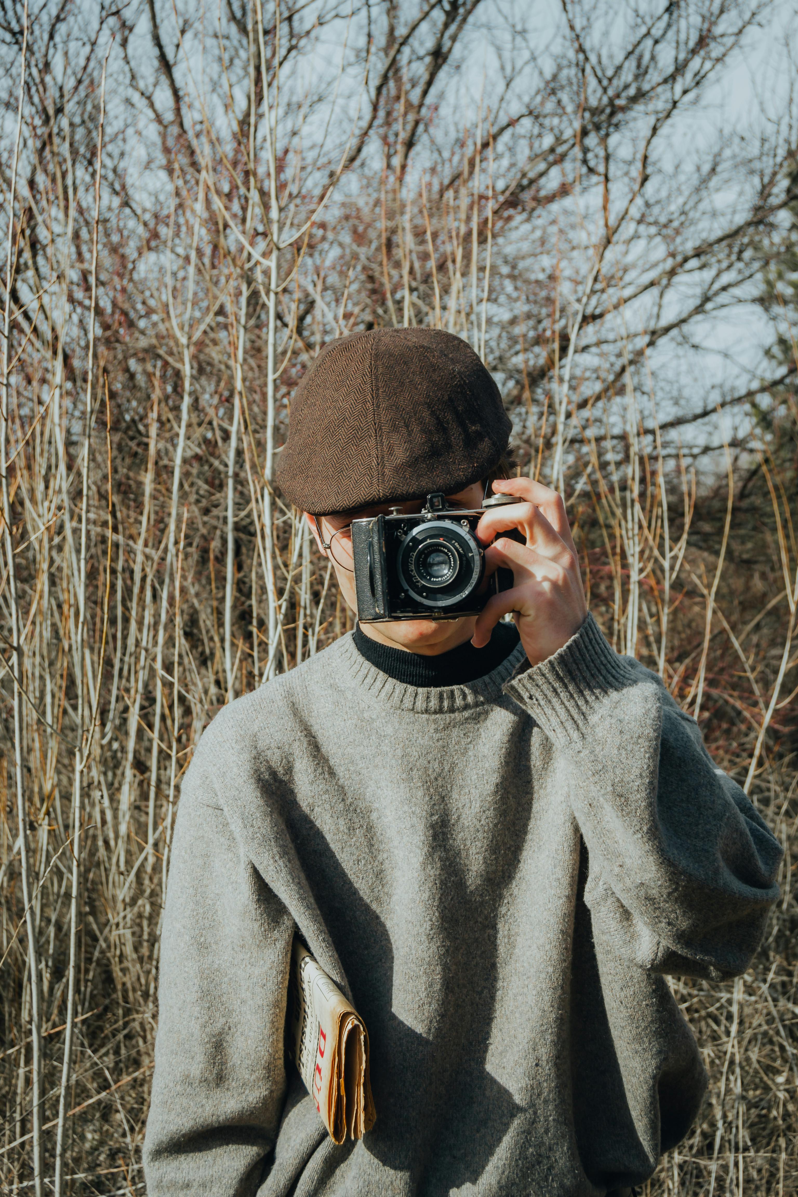 Free Man in a grey sweater and cap capturing images with a vintage camera outdoors. Stock Photo