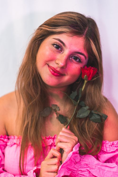 Free Smiling young woman in a pink blouse holding a red rose, conveying warmth and happiness. Stock Photo