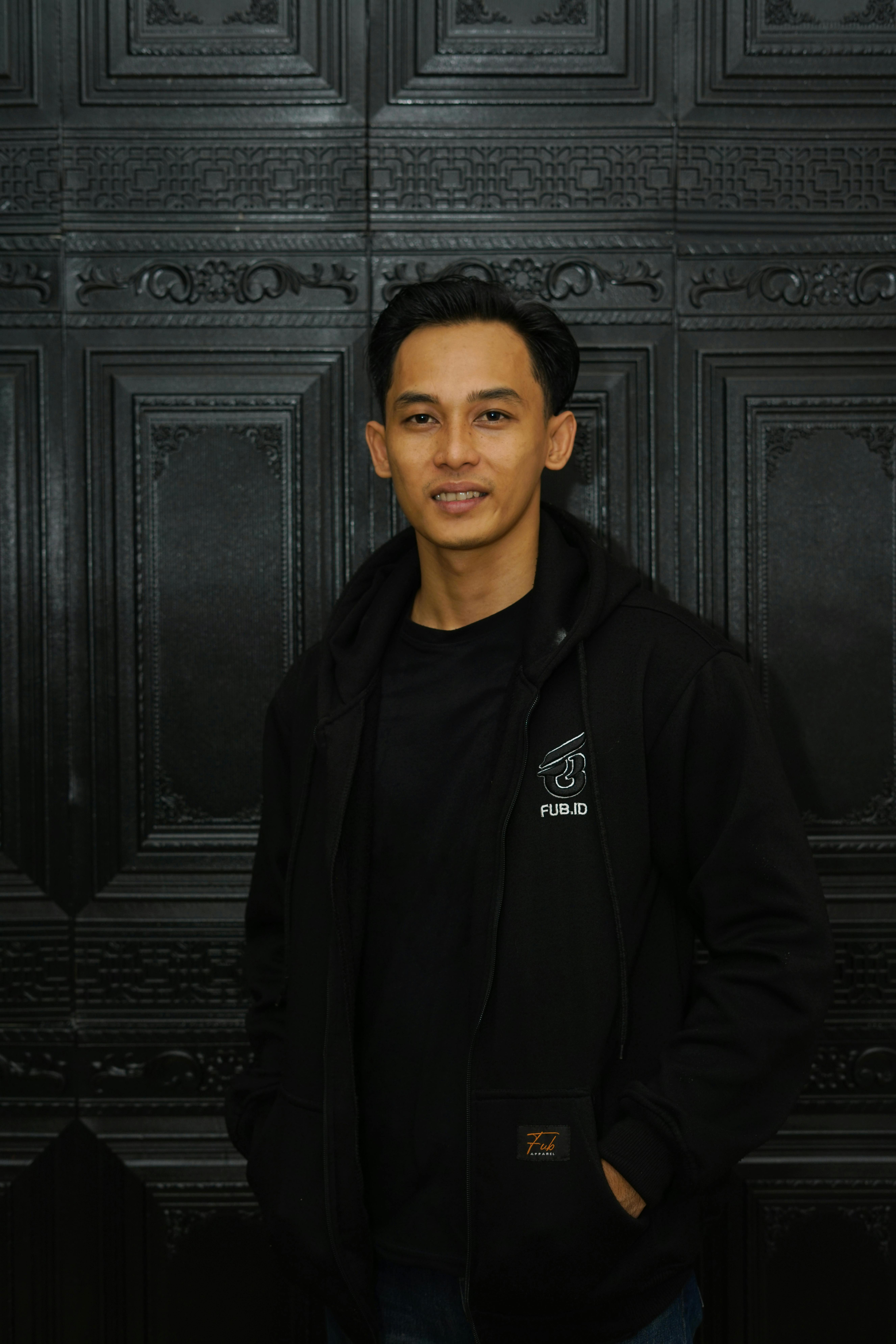 A man wearing a black hoodie poses confidently in front of a textured, ornate black wall.