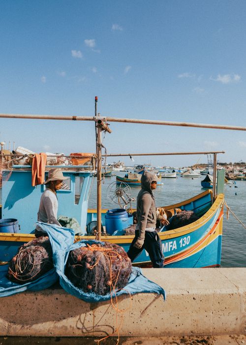Free Fishermen at Marsaxlokk harbor with colorful boats and nets, under a clear blue sky. Stock Photo