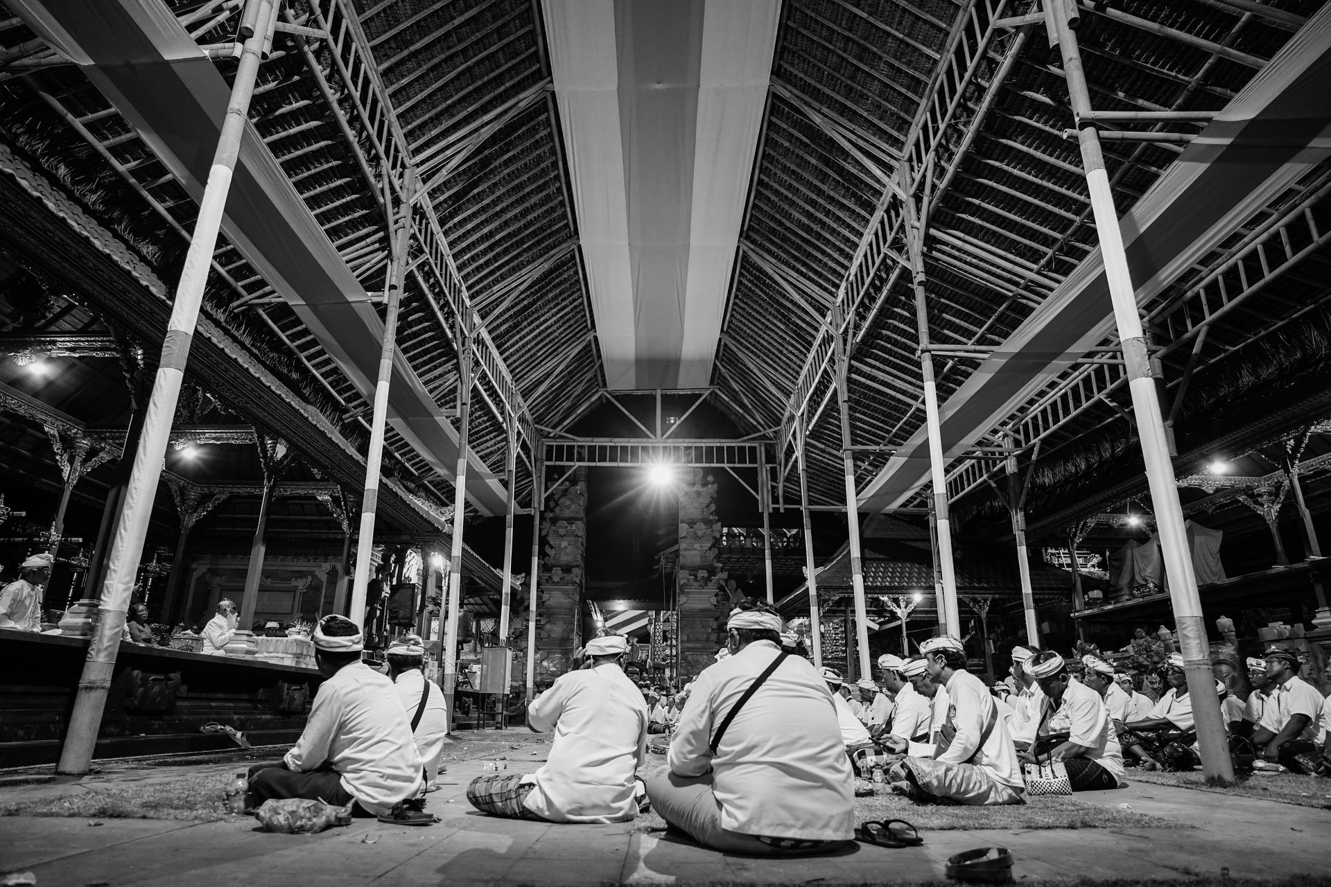 A gathering of men in traditional dress attending a night ceremony in a spacious open hall.
