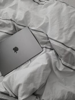 A modern laptop resting on soft, rumpled white and gray bedding, creating a minimalist and relaxed setting.