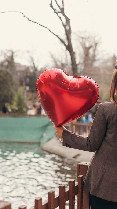 A Woman Holding a Heart Shaped Balloon Standing near a Fence 