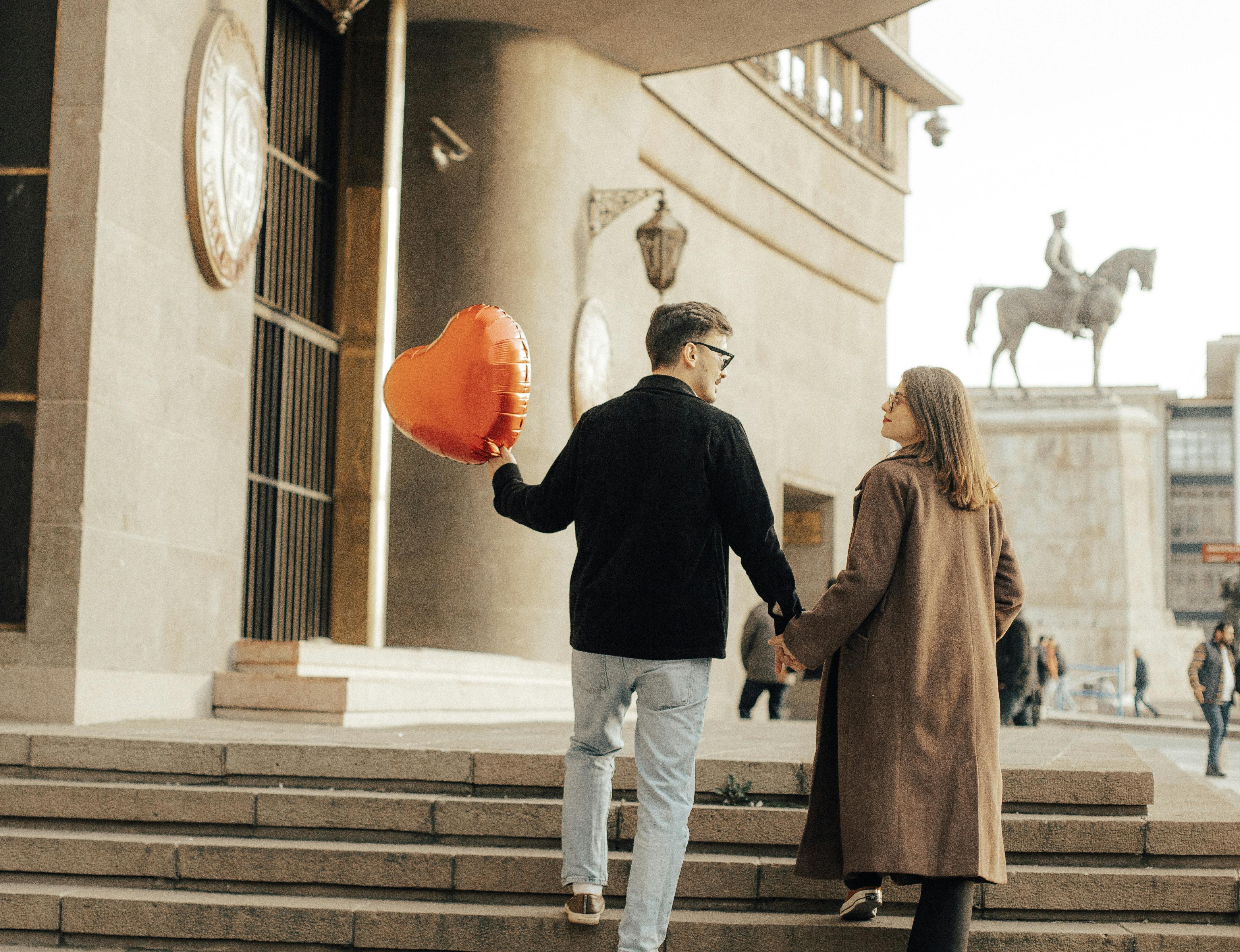 Couple holding hands with a heart-shaped balloon, walking up city steps. Urban romance captured outdoors.