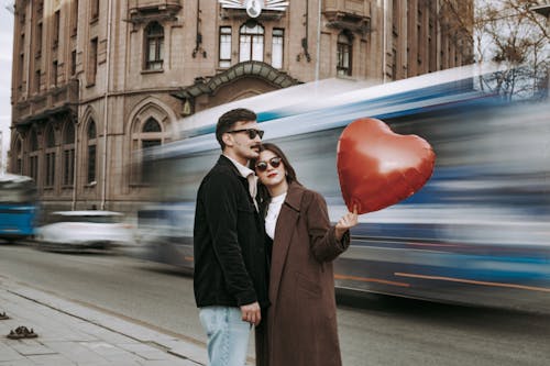 A Couple Holding a Heart Shaped Balloon Standing near a Street in City 
