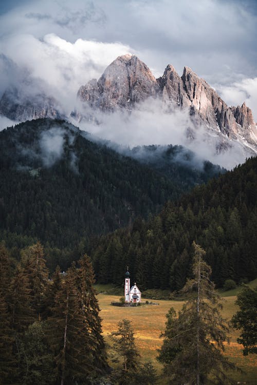 Little Church peeking thro forest in front of cloudy mountains