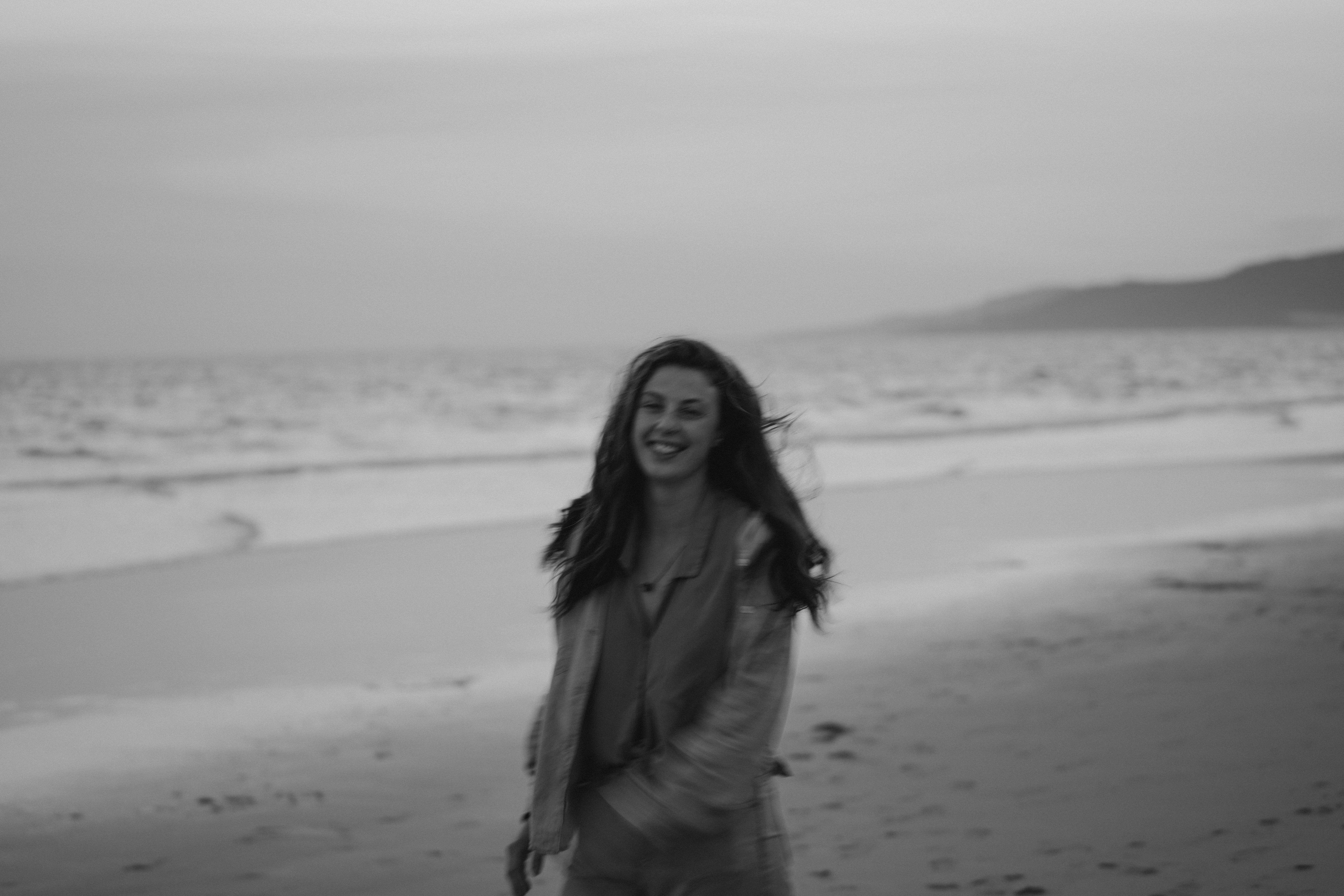 A woman joyfully walks on a sandy beach with waves in the background, captured in black and white.