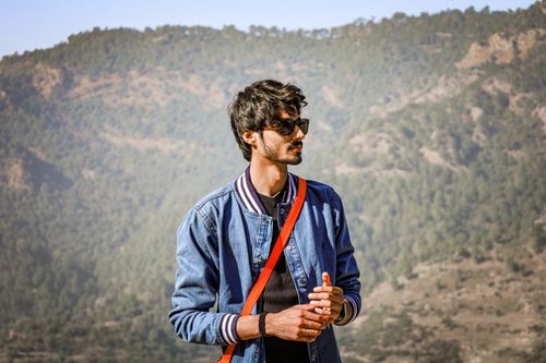 Free Man wearing sunglasses and denim jacket enjoying a scenic mountain view in summer sunlight. Stock Photo