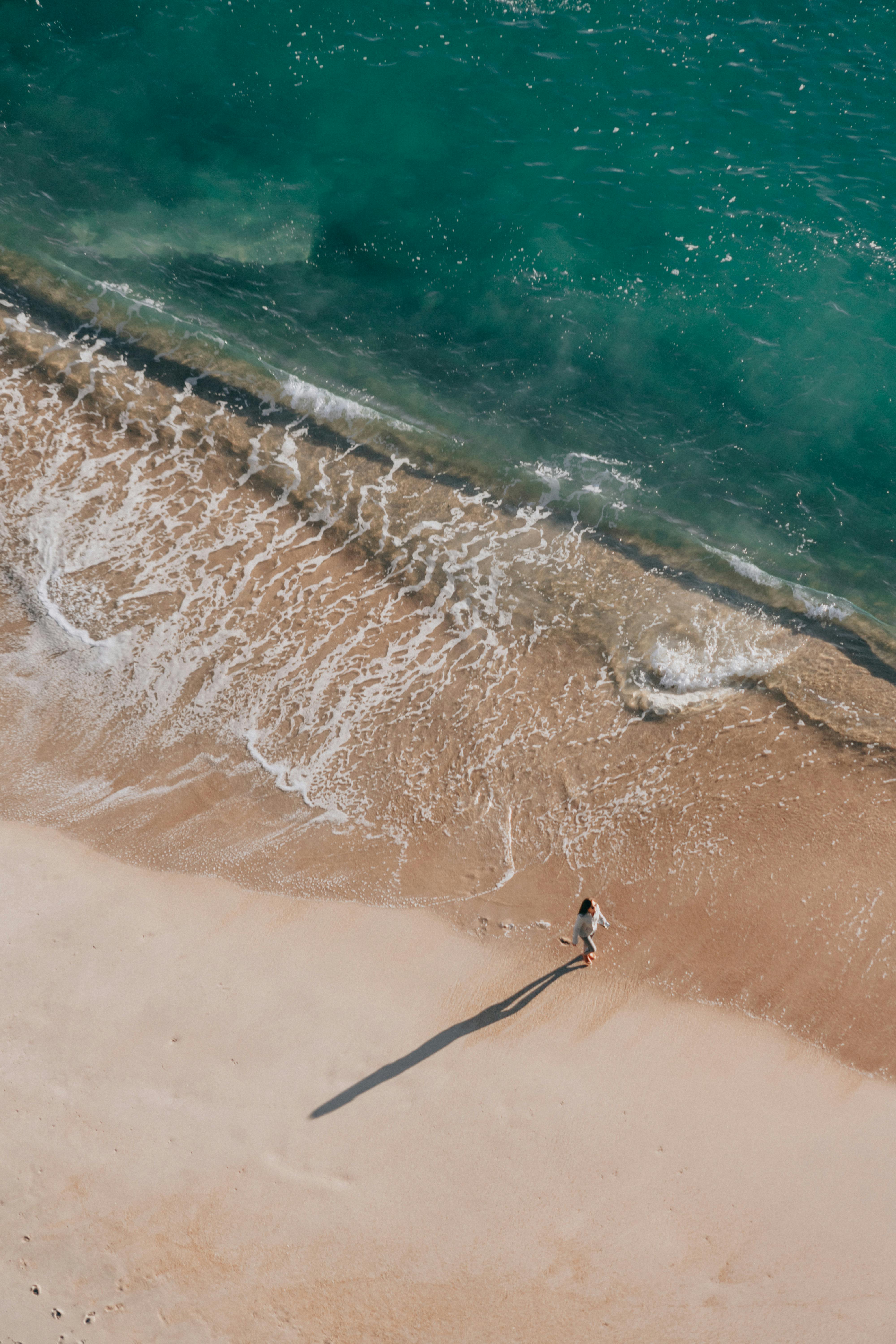 Aerial view of a woman walking along a tranquil sunlit beach with gentle waves.