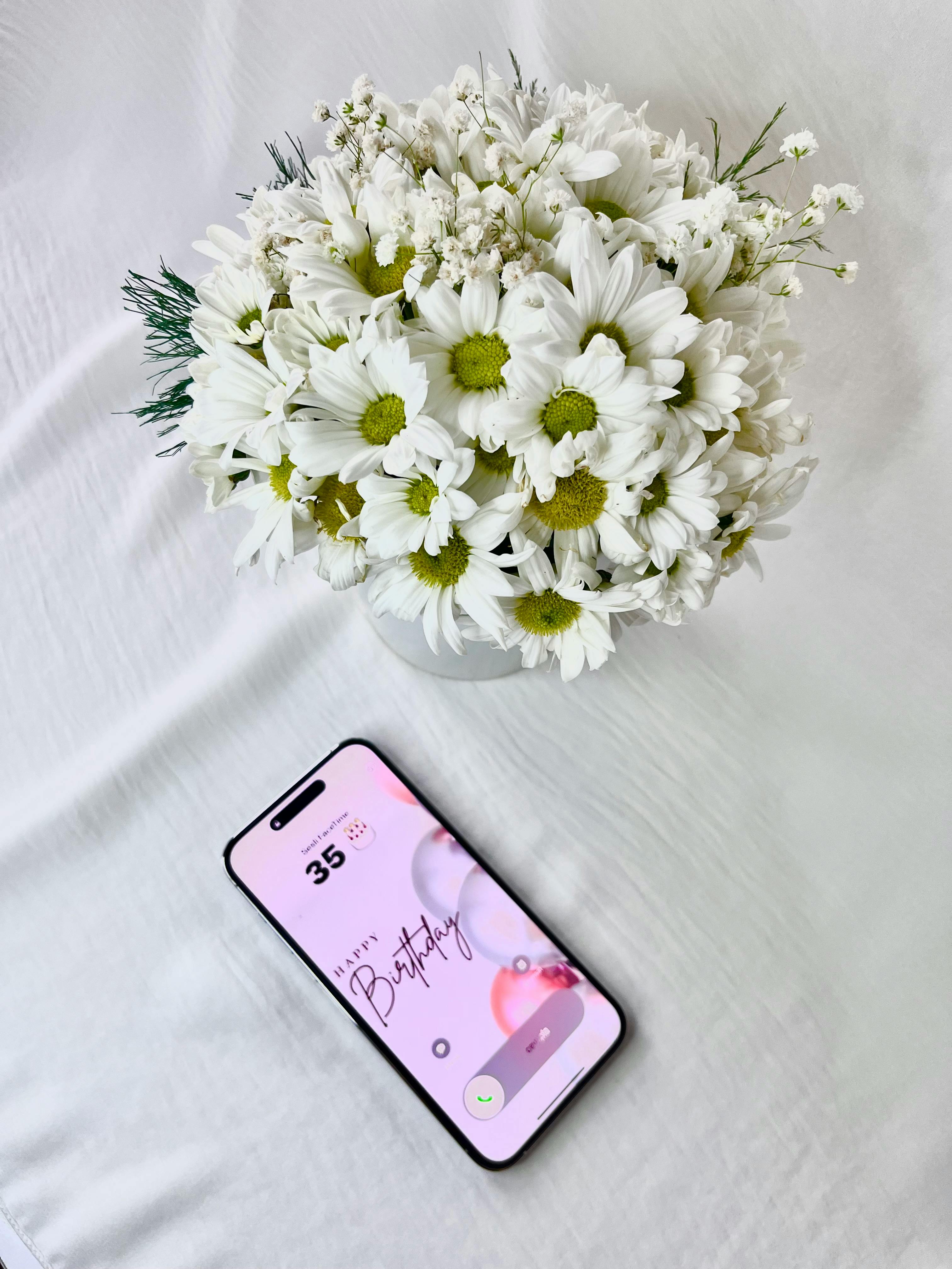 Vibrant white daisies in a vase beside a smartphone displaying a birthday message on a white background.