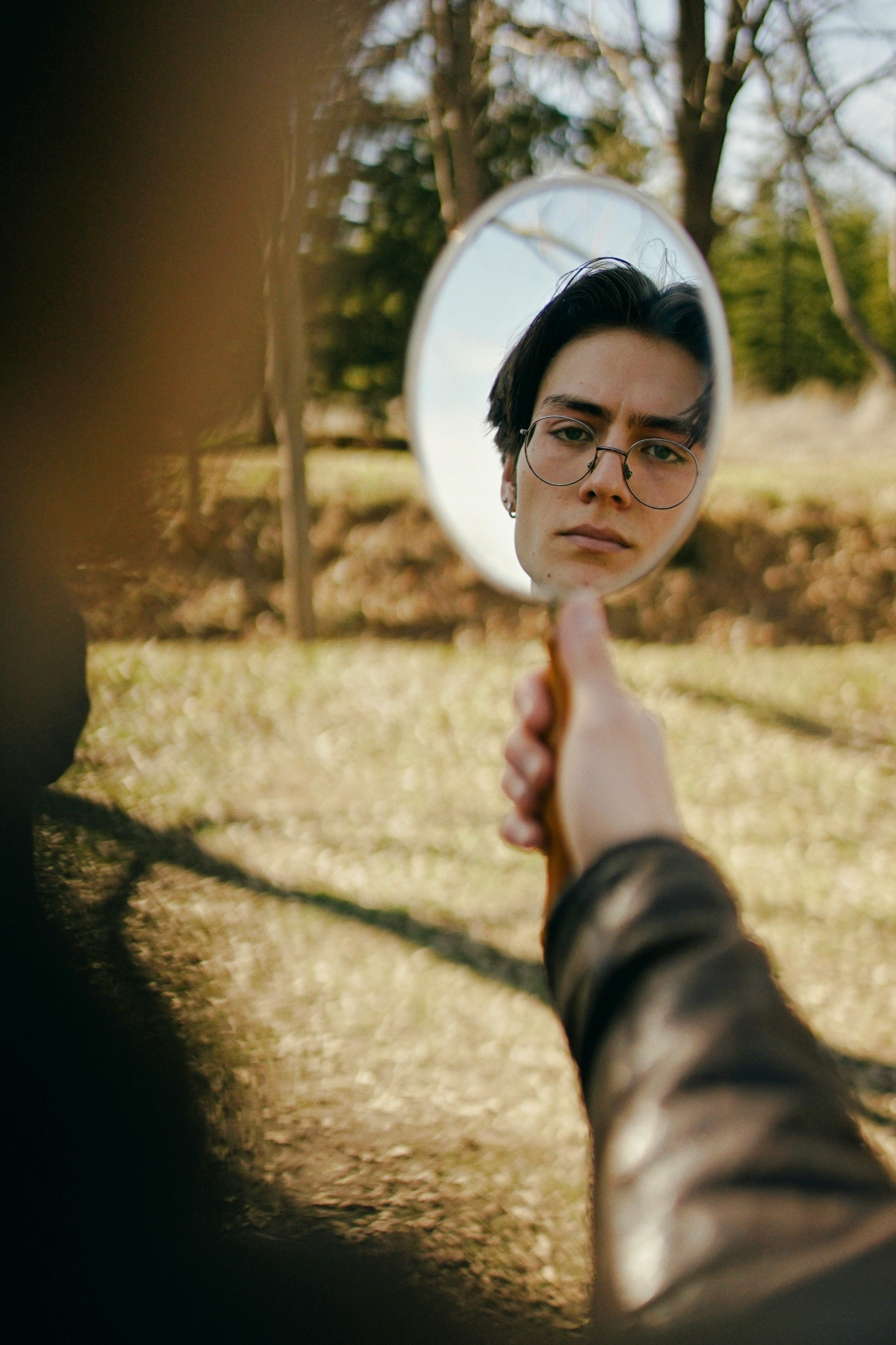 A man holds a mirror reflecting his face amidst an autumn park setting, creating a contemplative and artistic scene.