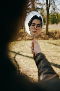 A contemplative young man gazing into a handheld mirror in an outdoor setting.