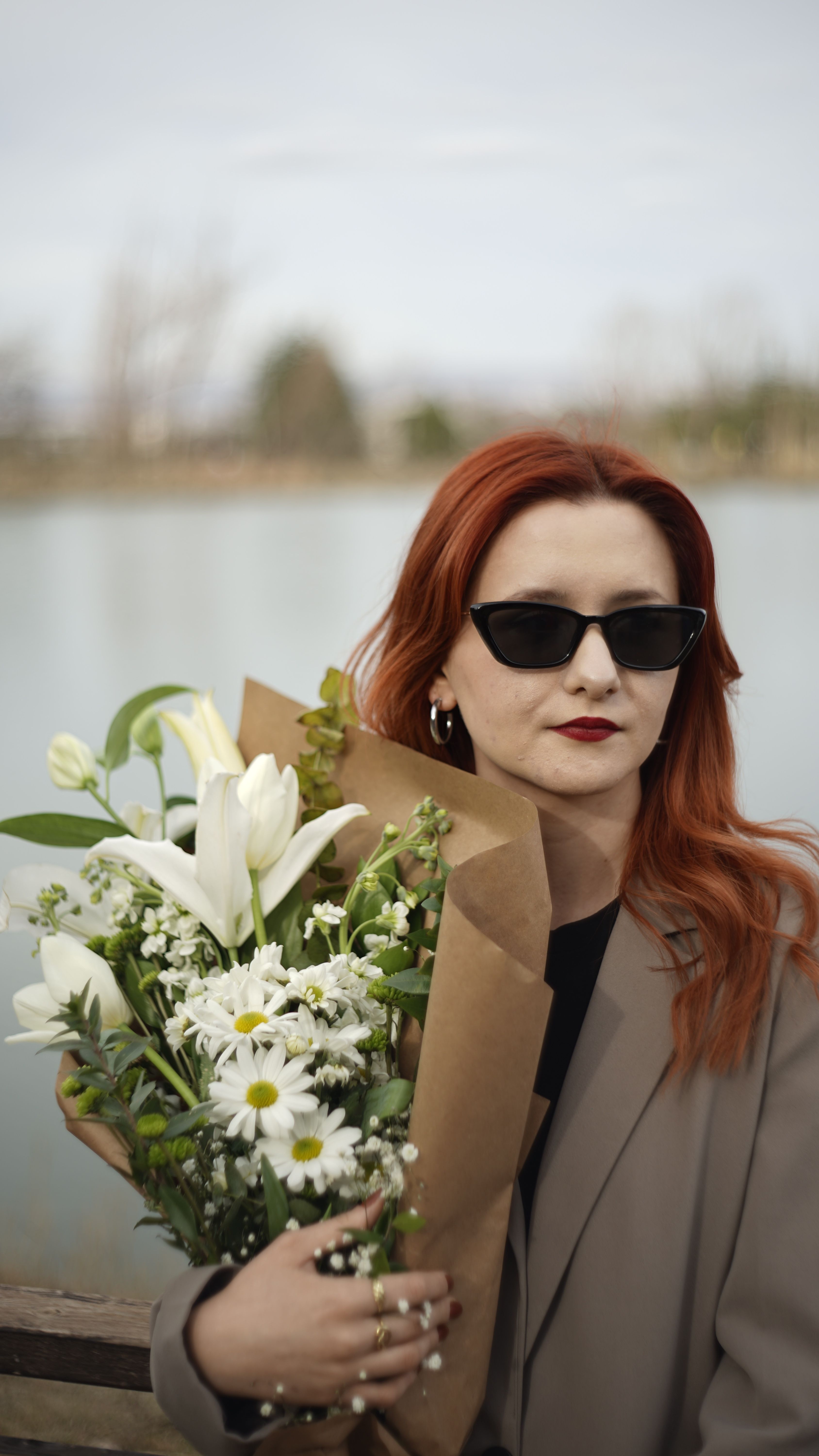 Elegant woman in sunglasses holding flowers by a tranquil lake