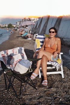 A woman in a swimsuit enjoys a sunny day on a pebbled beach, relaxing in a chair with a book.