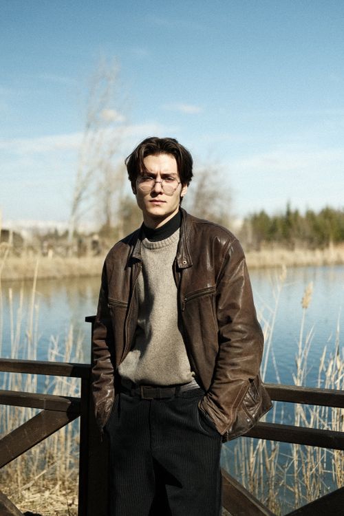 Free A stylish young man in a leather jacket poses on a footbridge by a river during fall. Stock Photo