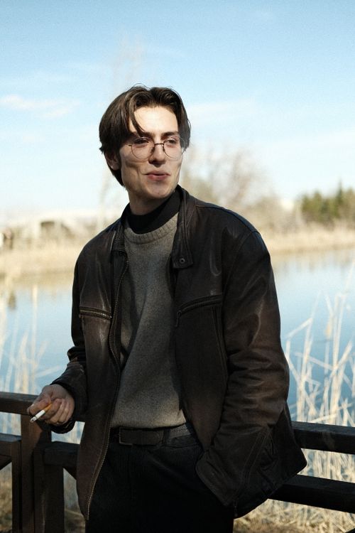 Free Stylish man in a leather jacket enjoys a cigarette by a river with a bridge in the distance. Stock Photo