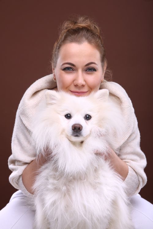 Free A woman smiles warmly while holding her fluffy white Japanese Spitz dog against a brown background. Stock Photo