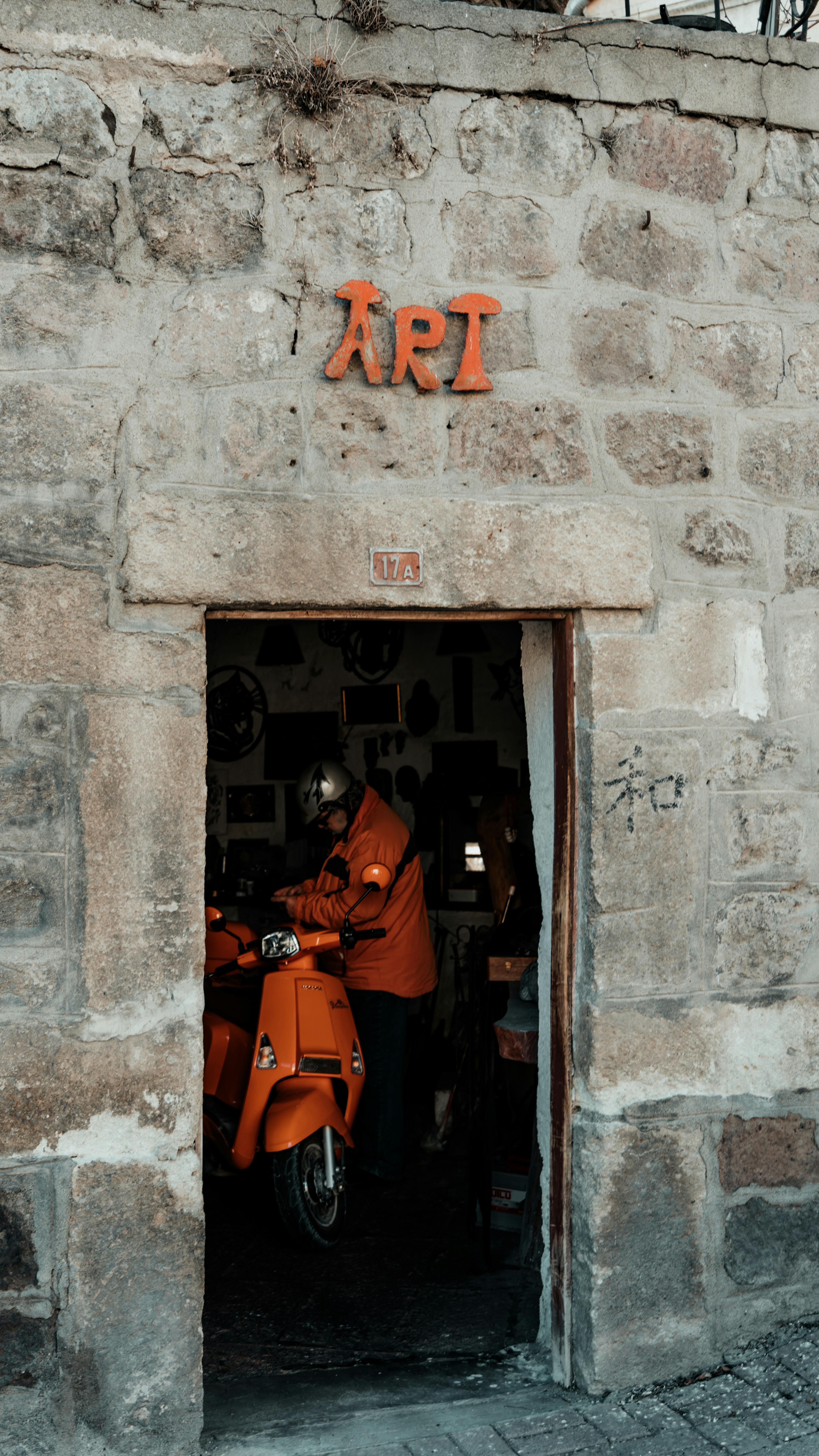 A deliveryman in an orange jacket enters an old stone building with a scooter, emphasizing urban life.