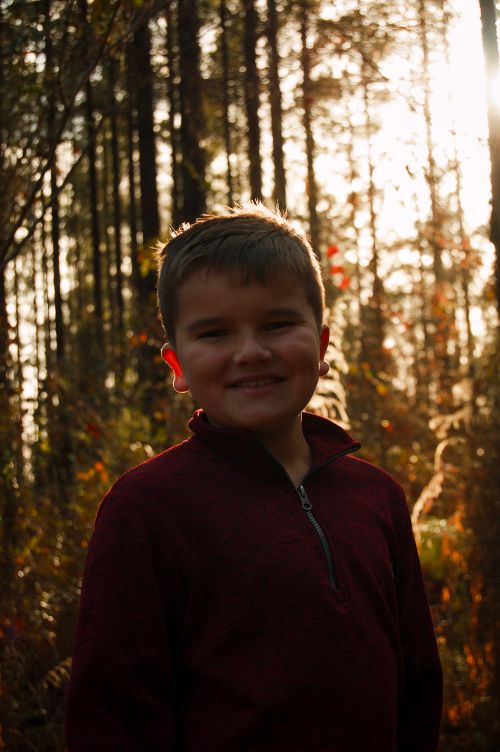 Free A cheerful young boy with short hair smiling brightly against a sunlit forest backdrop. Stock Photo