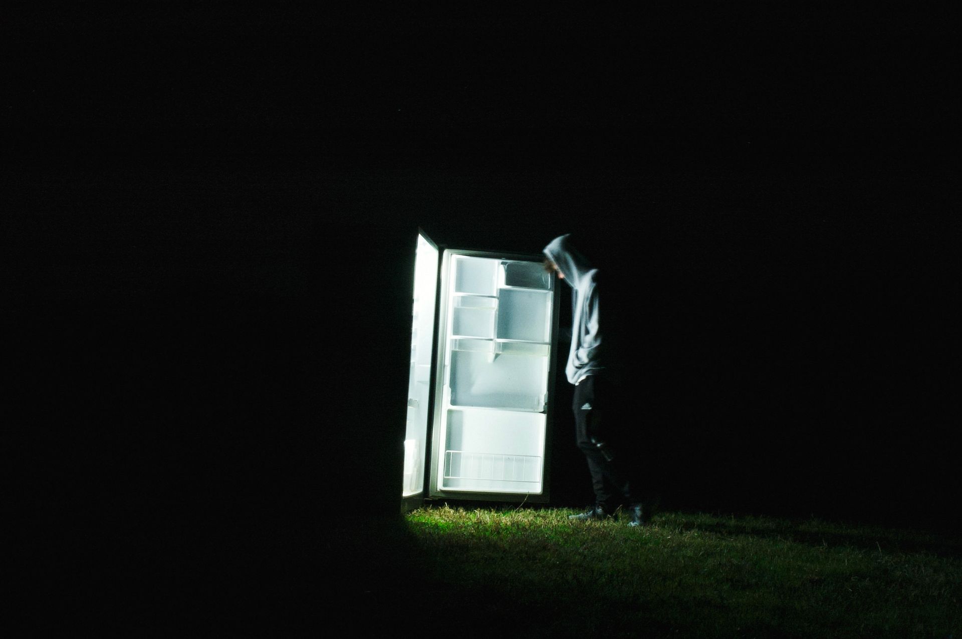 A person in a hoodie stands by an open fridge in the dark night, creating an eerie atmosphere.