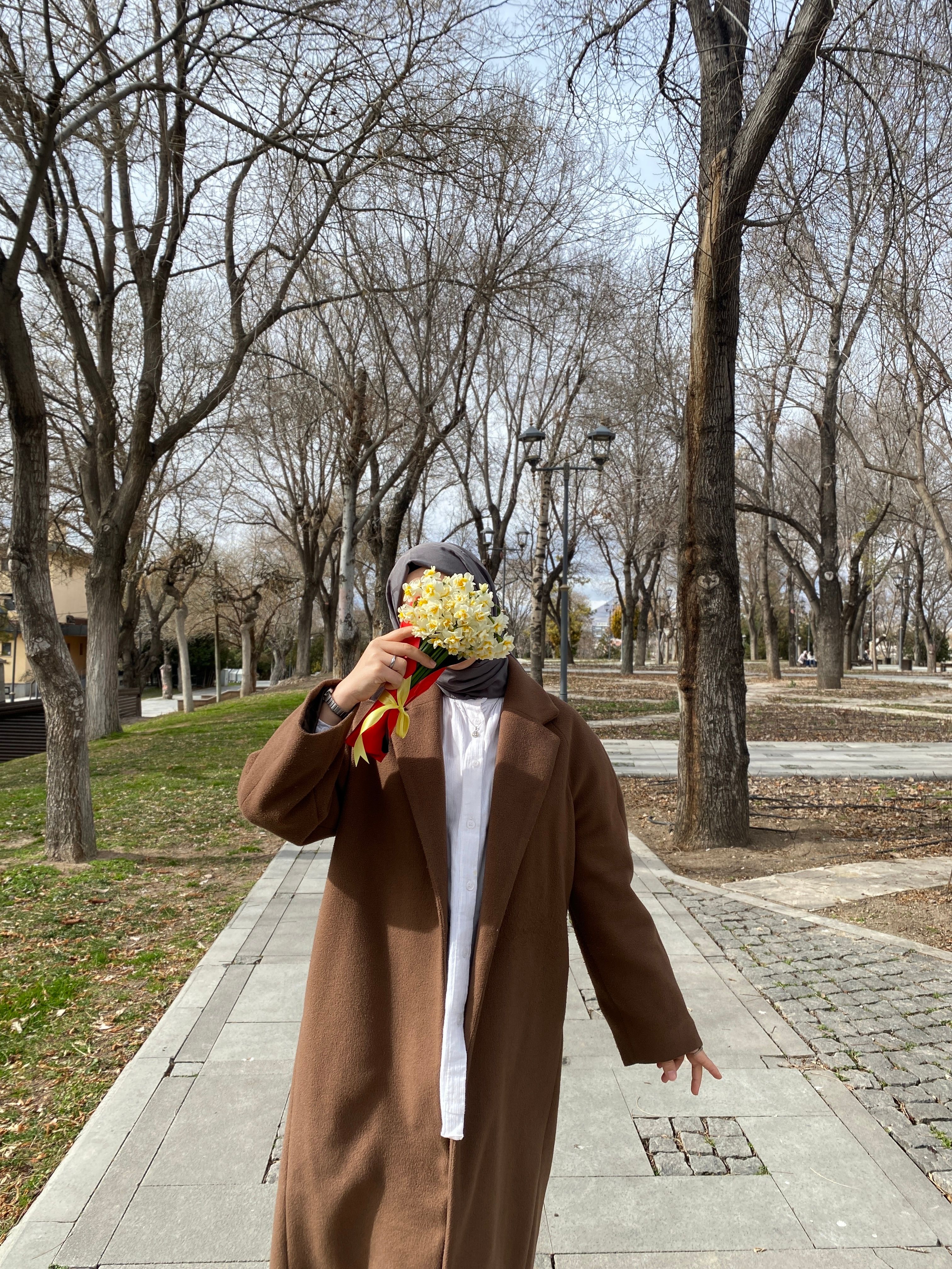 A woman wearing a hijab and brown coat holds flowers in an urban park.