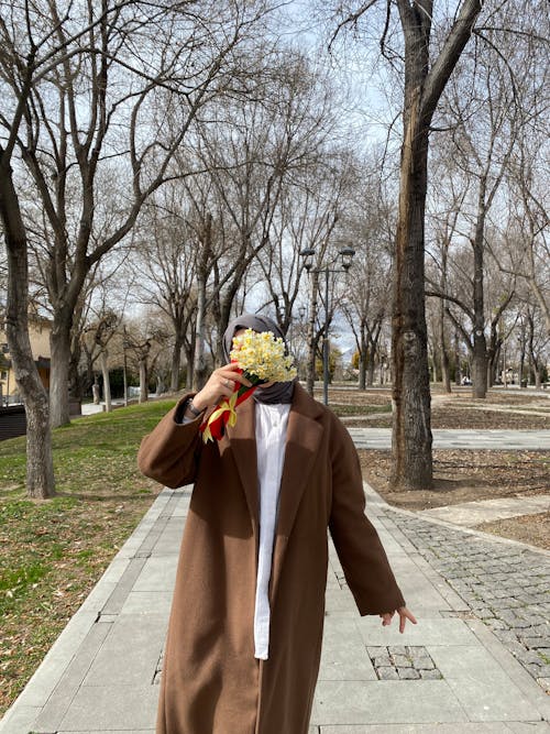 Woman in Brown Coat and Hijab Holding Flowers and Standing at Park