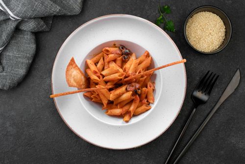 Free Top view of a penne pasta dish with olives, sauce, and a crispy bread stick. Stock Photo