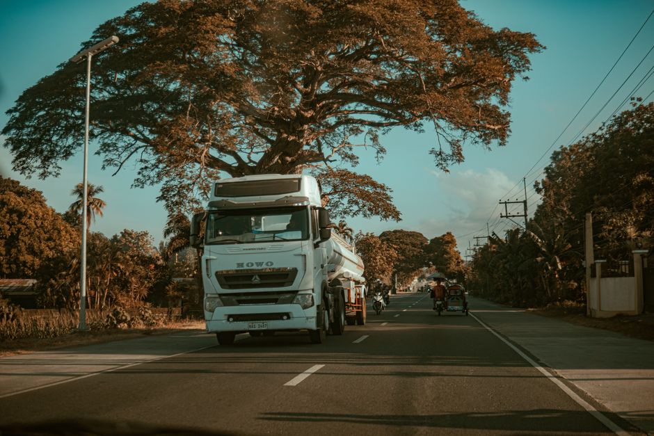 A large truck travels down a rural road lined with lush trees under a clear sky.