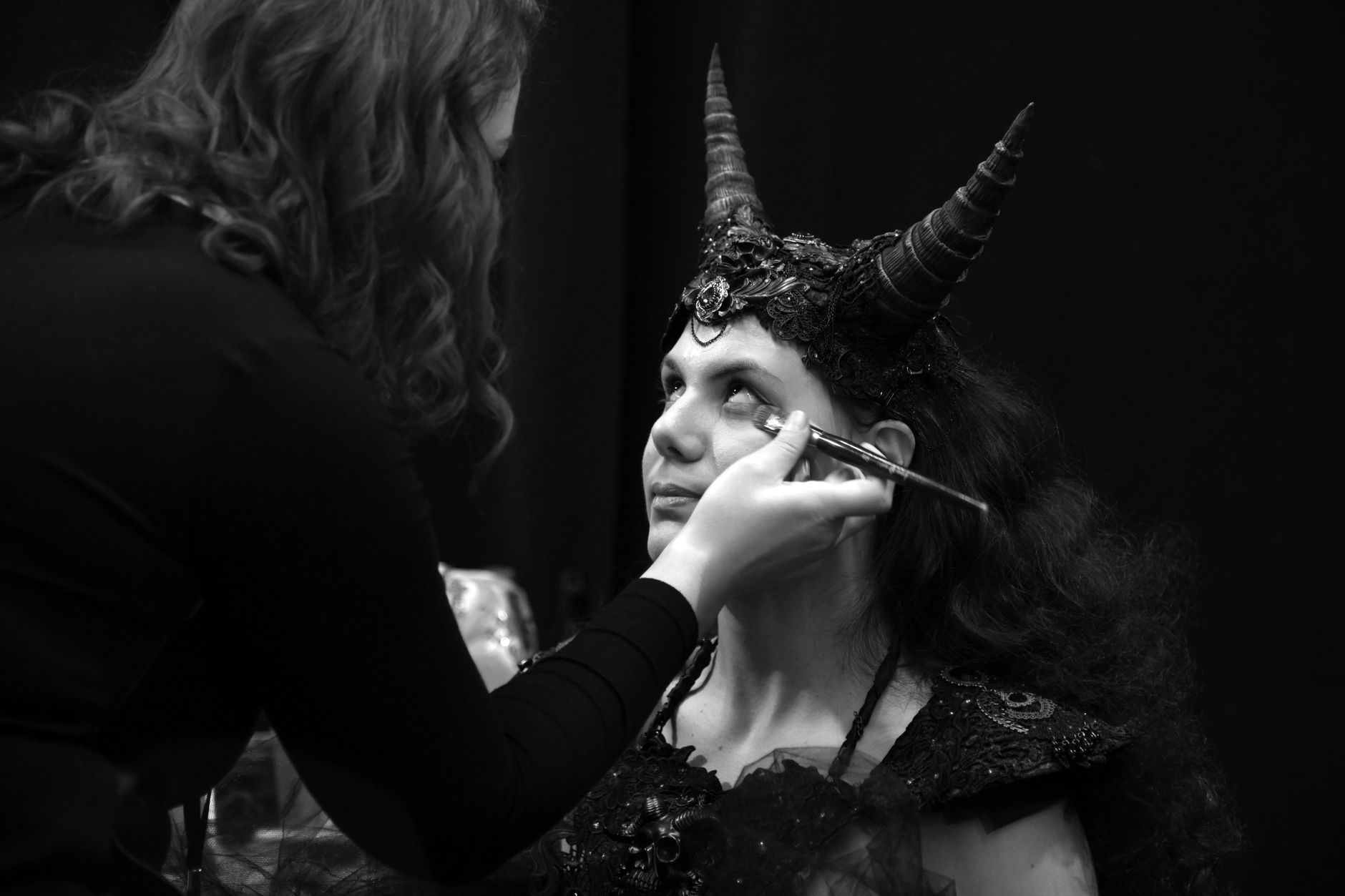 Black and white photo of a woman receiving makeup for an elaborate horned costume.