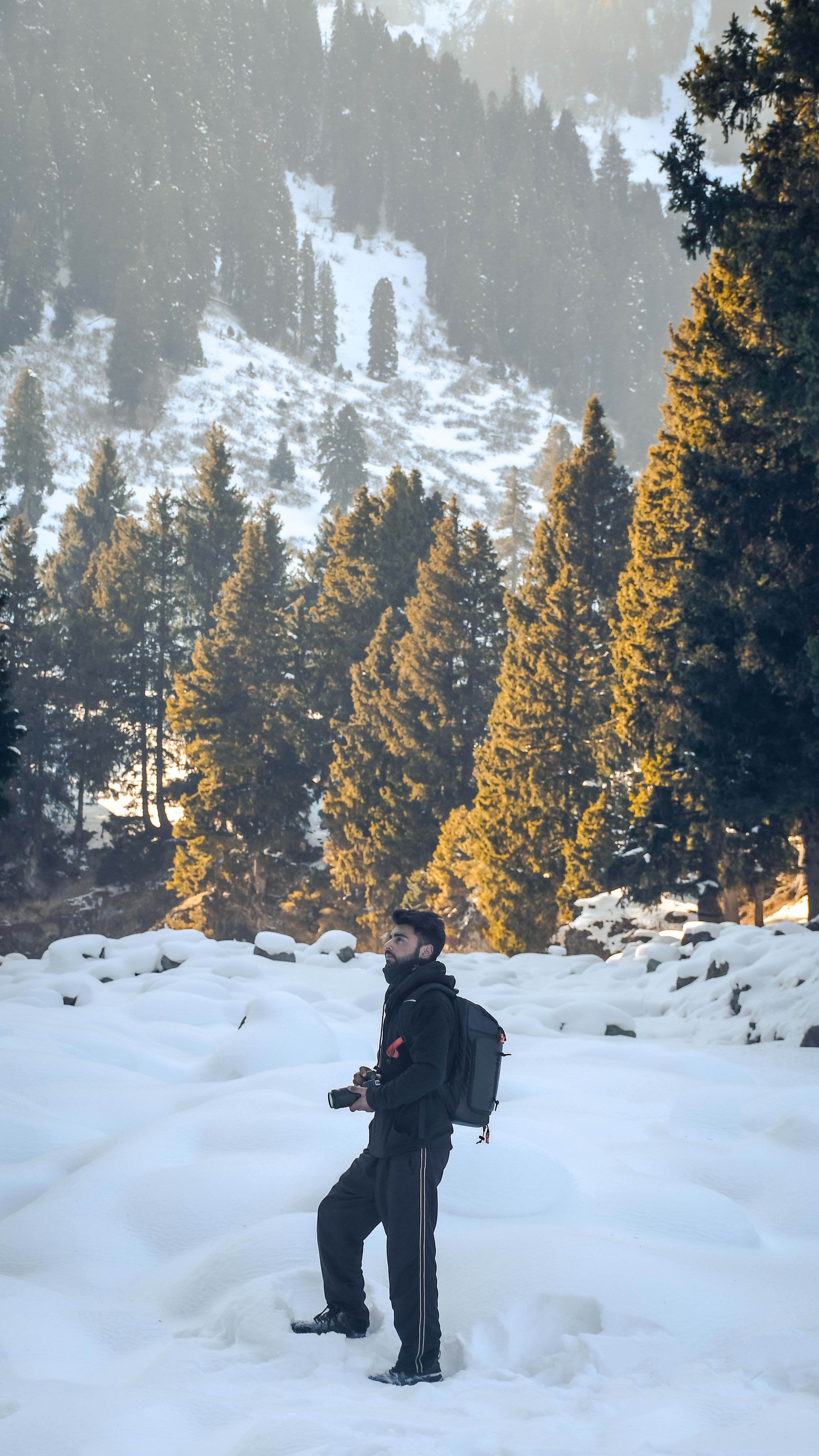 A man with a backpack and camera stands in a snowy forest during winter, capturing adventure moments.