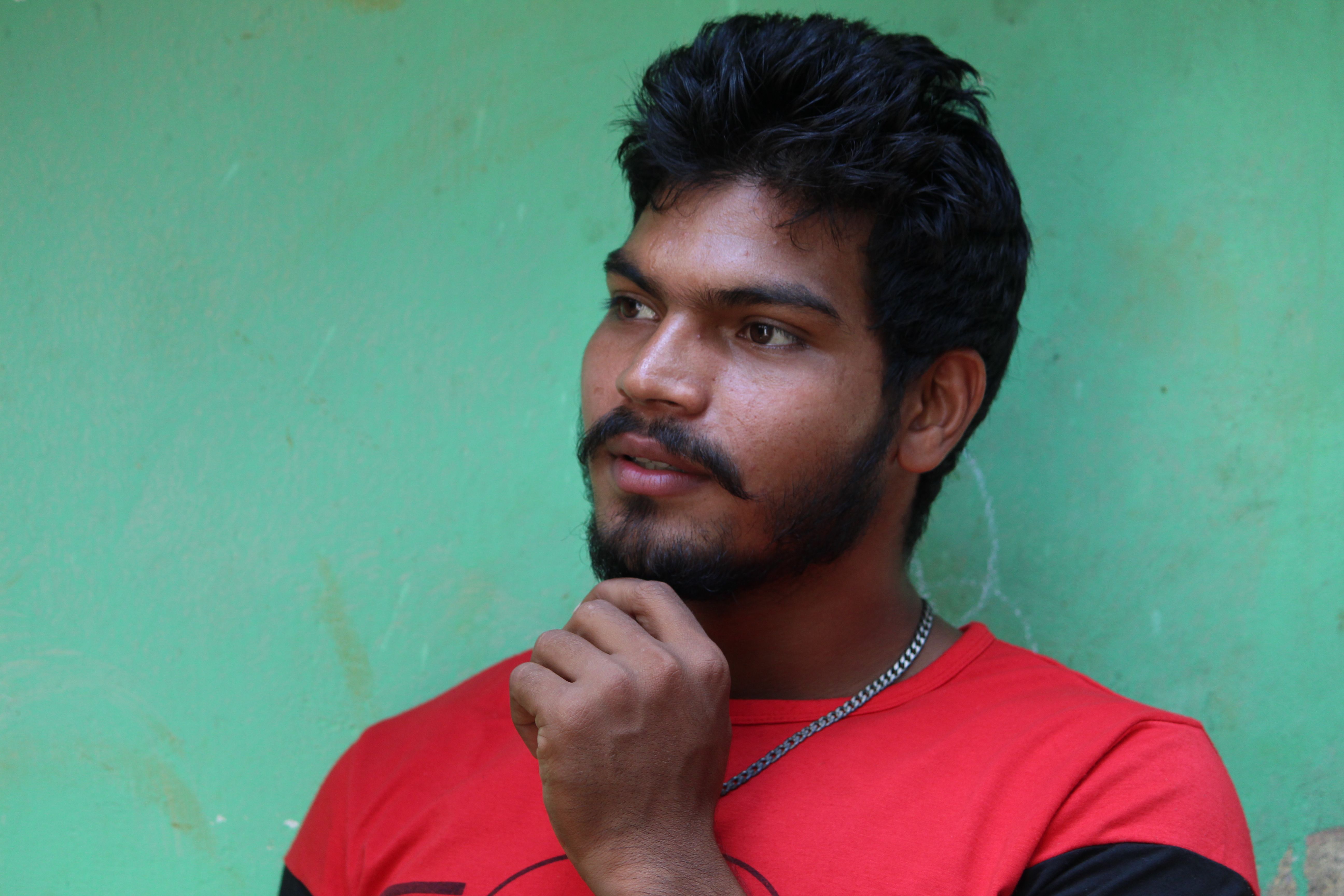 A thoughtful man with facial hair poses against a green wall, wearing a red shirt.