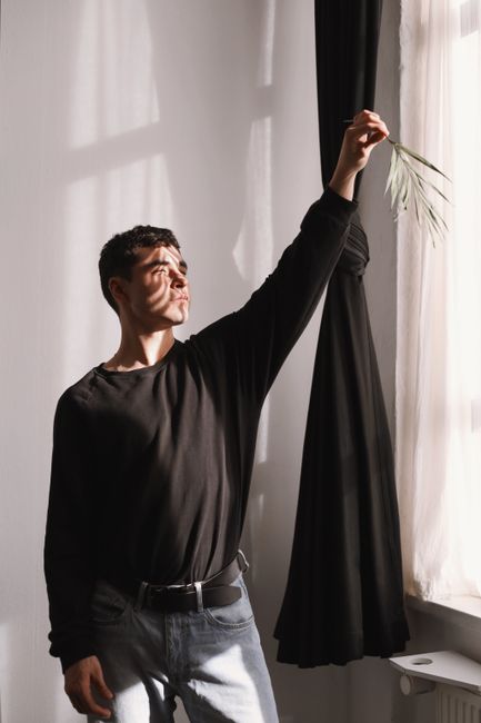 A man in casual attire stands by a window, holding a leaf, with light casting dramatic shadows.