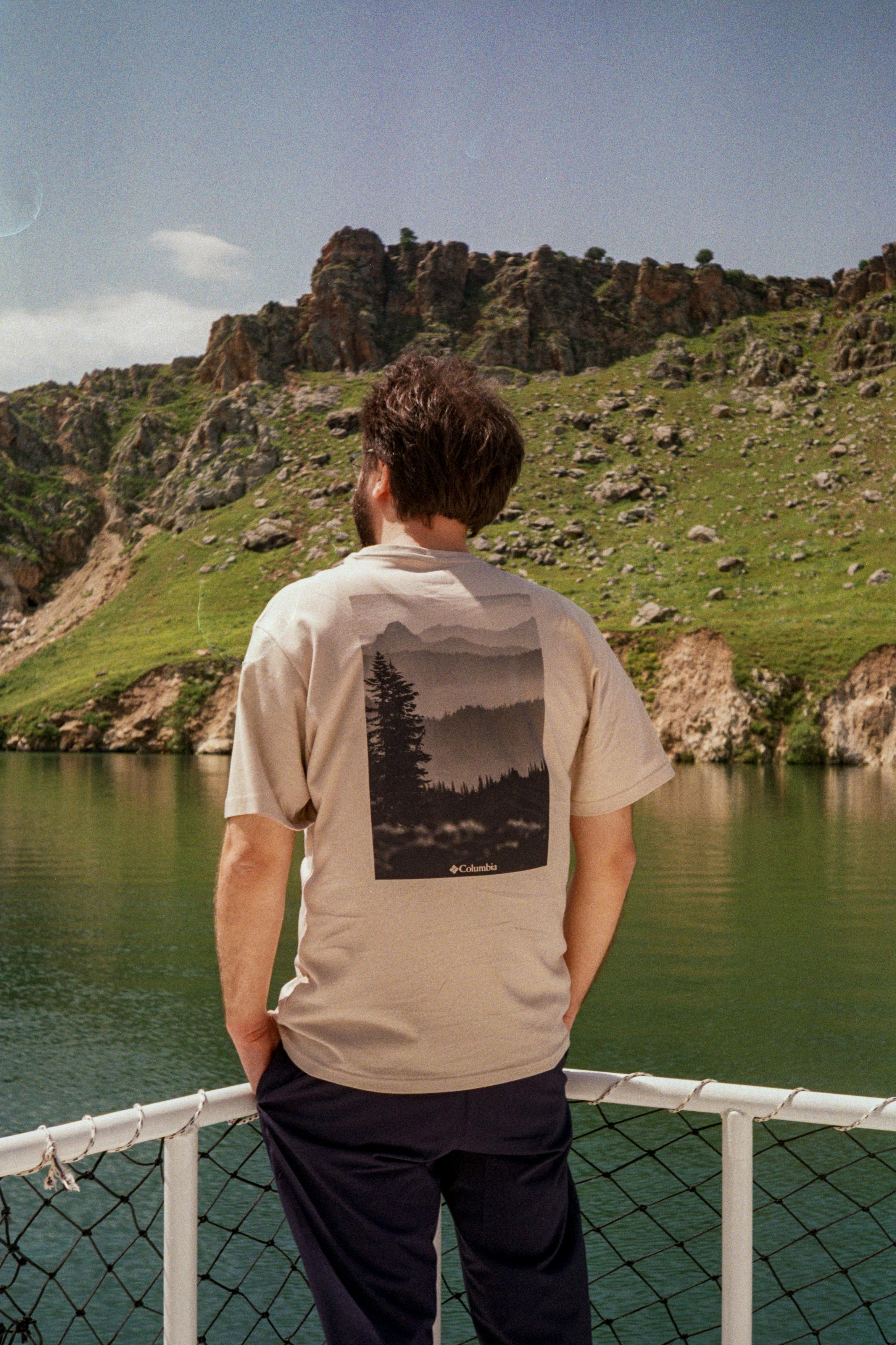 Man standing on a boat, overlooking a tranquil lake and lush mountains in summer.