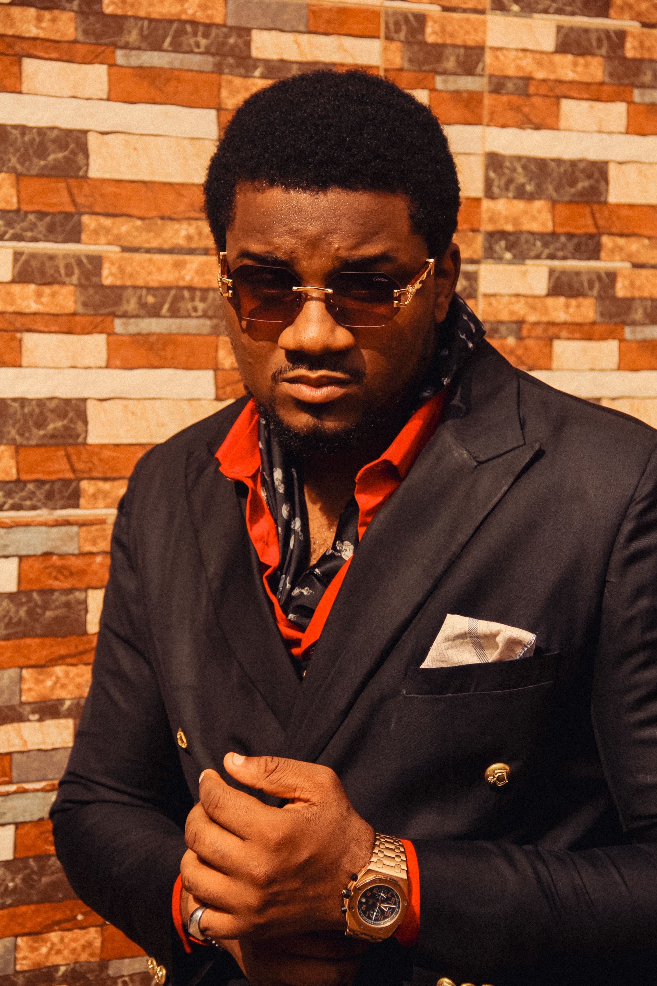 Confident man with sunglasses and watch in black suit against brick wall.