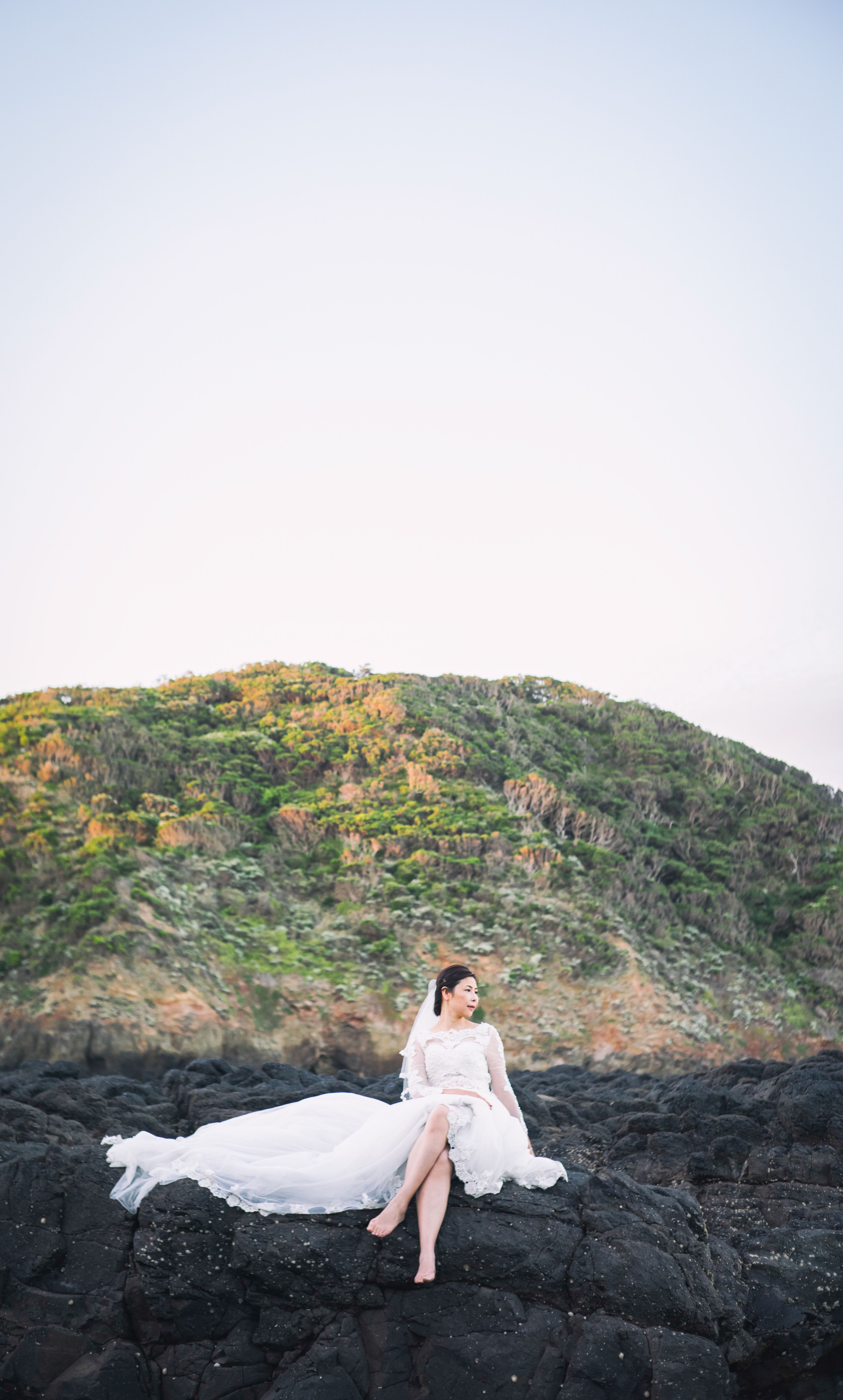 A bride in a white wedding dress sits elegantly on volcanic rocks against a mountain backdrop.