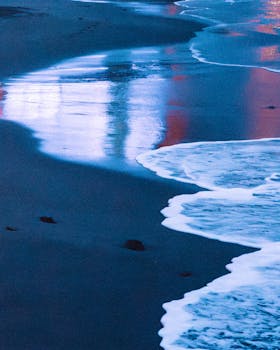 Twilight waves gently lap at the beach, reflecting serene blues and whites.