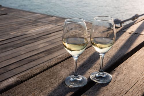 Free Elegant white wine glasses on a rustic wooden pier overlooking water at sunset. Stock Photo