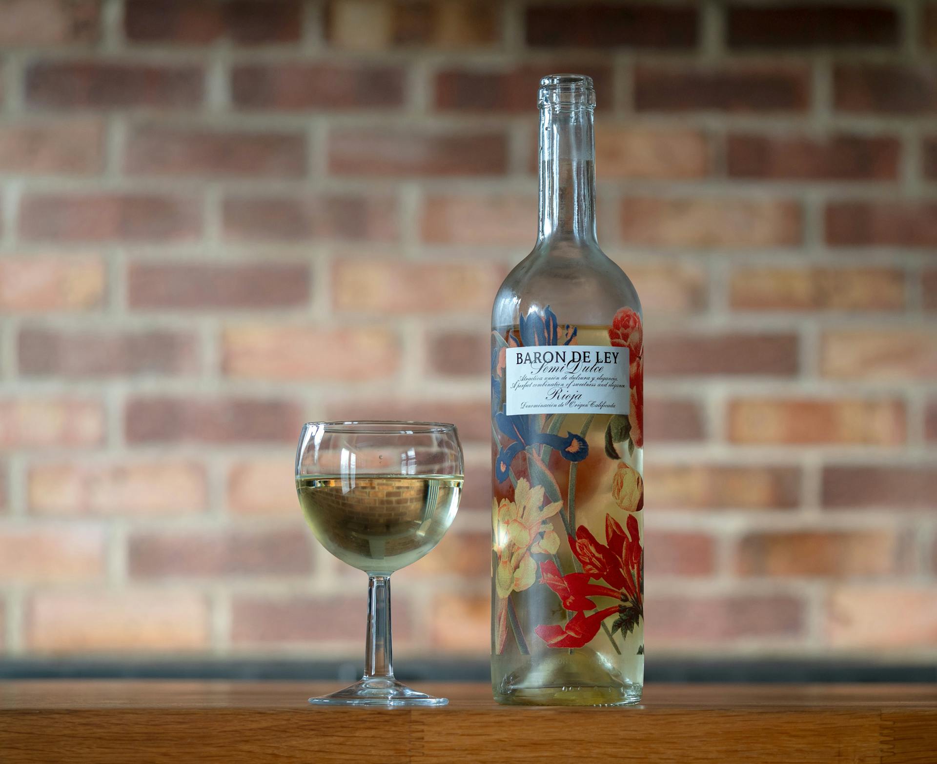 A Baron de Ley wine bottle and glass on a wooden table with a brick wall background.