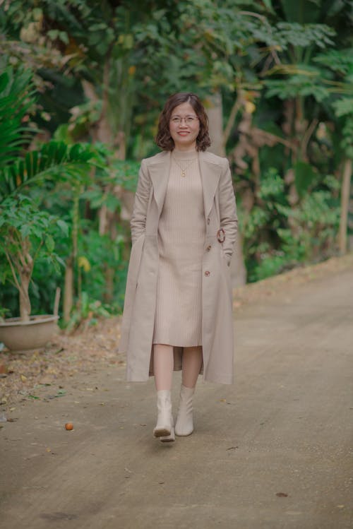 Woman in a Beige Trench Coat and Dress Walking Along the Road