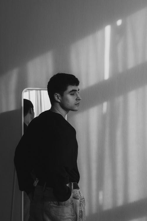 Black and White Photo of a Young Man Standing by the Mirror 