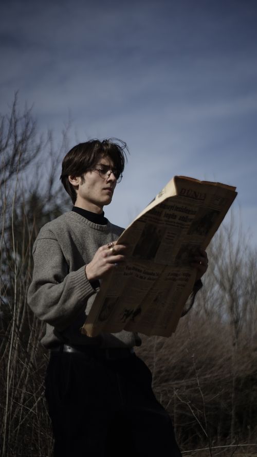 Free A young Asian man in a sweater reads a newspaper outside in a park in Ankara, Türkiye. Stock Photo