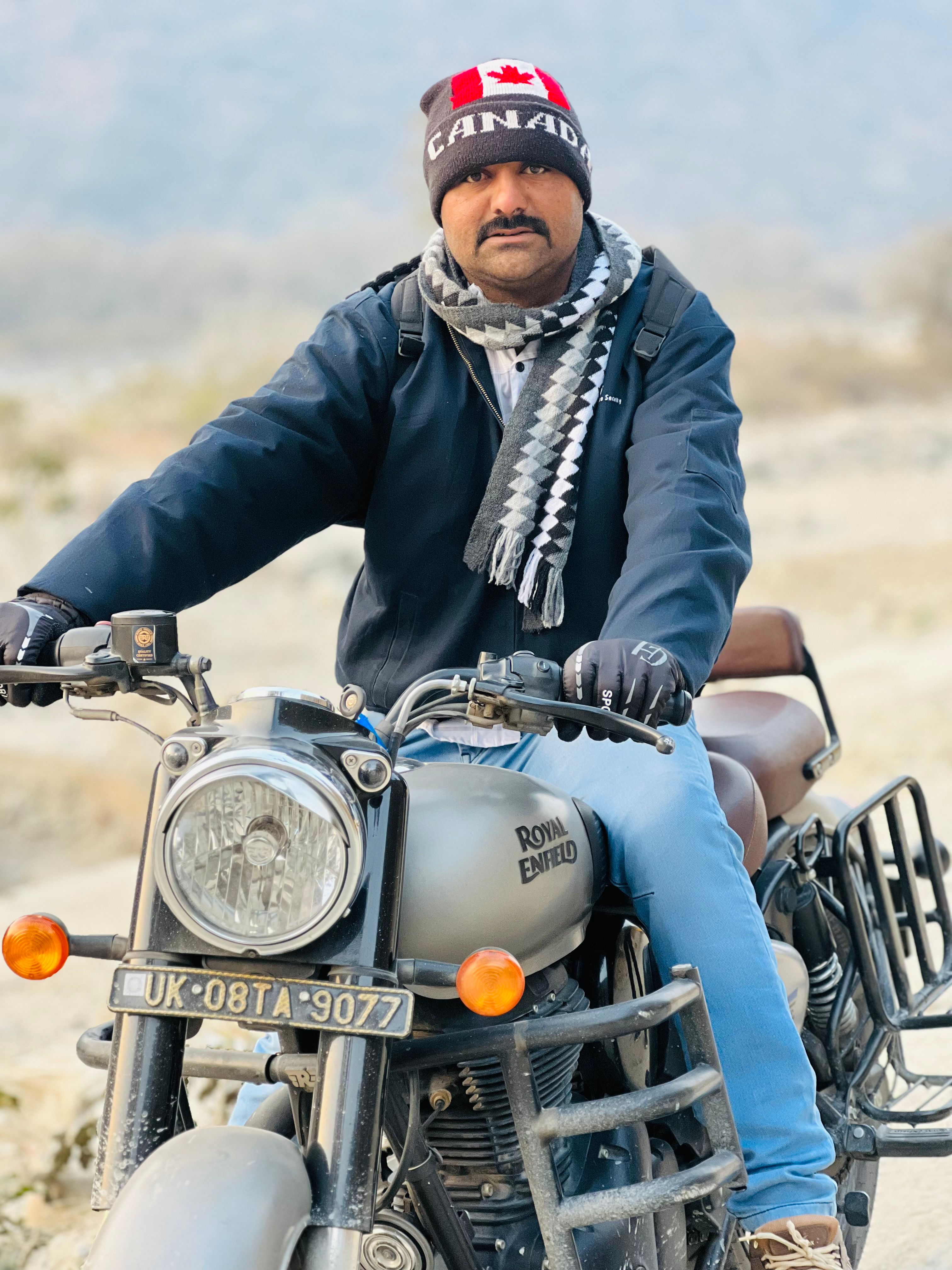 Portrait of a man with a Canada hat riding a Royal Enfield motorcycle in a rural setting.